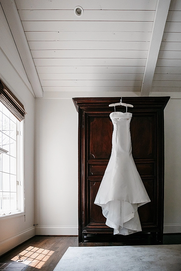Wedding dress hanging on a hanger, a strapless wedding dress in window light against a dark wood armoire in a bright room