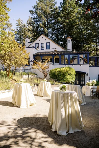 Wedding cocktail hour with ivory linen cocktail tables, floral centerpieces and candles under string lights on a sunny patio by a white stucco venue