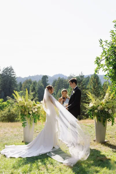 Wedding vows during an outdoor wedding ceremony as bride and groom hold hands, her long veil blowing, with mountains and pines behind