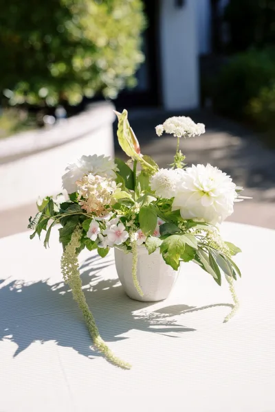 Wedding centerpiece with white floral centerpiece in a ceramic vase and greenery on a tablecloth, set on a sunlit outdoor patio with garden foliage