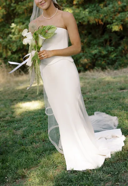 Bridal portrait of a bride in a strapless wedding dress with veil and pearl necklace, holding calla lilies and anthurium bouquet on a grassy lawn
