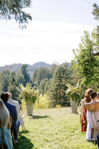 Outdoor ceremony setup with mountain wedding ceremony chairs lining an aisle, large white floral urns and greenery on a grassy lawn with trees and mountains