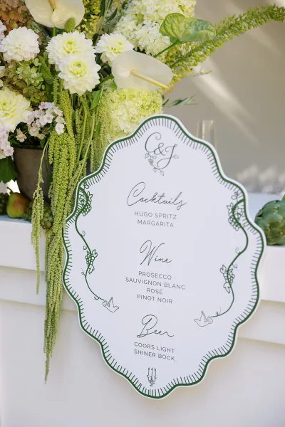 Wedding bar menu and signature cocktail sign on a white counter beside a white-and-green floral arrangement with anthurium and fruit accents