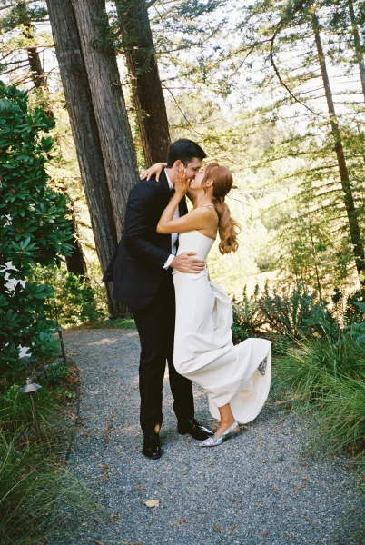 Wedding kiss as groom lifts bride in a strapless dress on a gravel path, forest trees and greenery behind, bride in silver flats