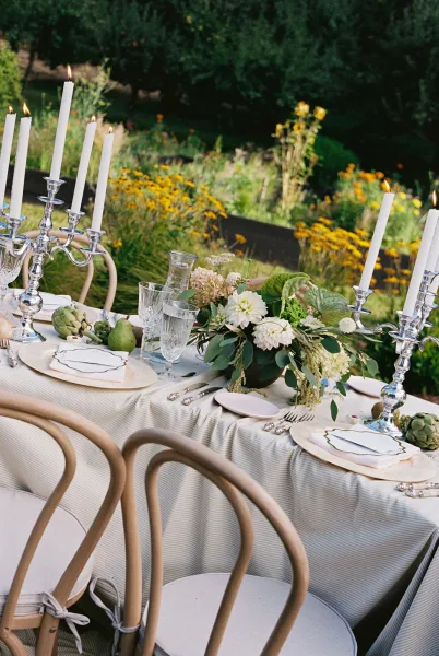 Reception tablescape with striped tablecloth, white taper candles in silver candelabras, and green-white florals on an outdoor reception table in a garden lawn