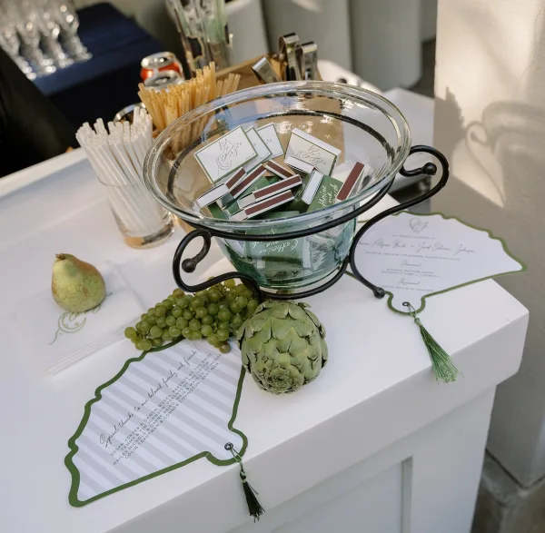 Wedding matchbook favors in a glass bowl on a metal stand with personalized matchbooks, straws, stirrers, and fruit on a white bar table