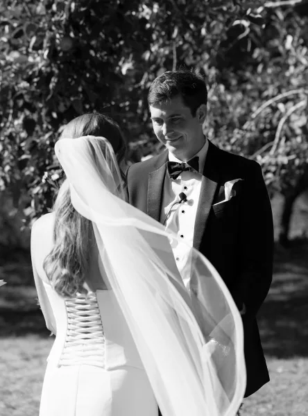 Wedding vows during an outdoor wedding ceremony as groom in tuxedo smiles at bride in long veil, with garden trees and lawn behind