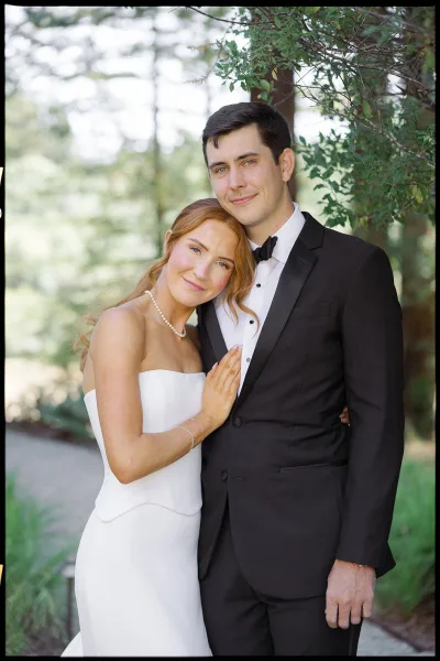 Couple portrait of bride in a strapless dress with pearl necklace leaning on groom in black tuxedo on a tree-lined outdoor path
