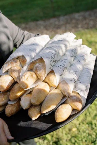 Wedding bread basket with wedding dinner rolls in paper cones on a serving tray, held by a server on a lawn beside a gravel path