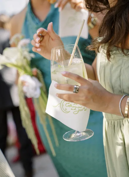 Signature cocktail in a clear glass with mint garnish and paper straw, held by a guest in a green dress with a bouquet behind
