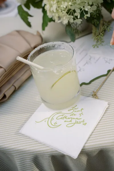 Wedding signature drink with signature cocktail sign, salted rim glass and lime wedge on striped tablecloth beside place cards and florals