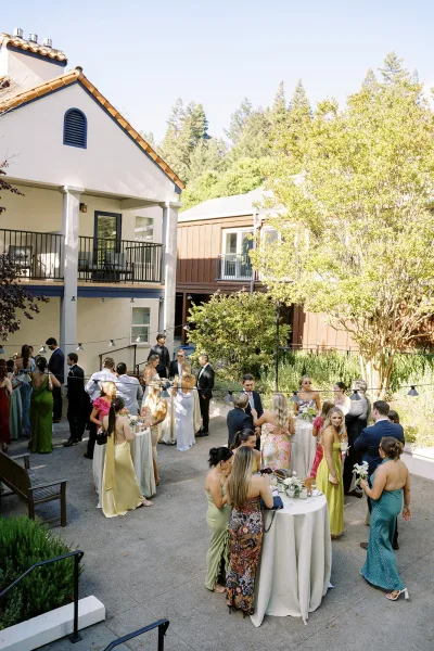 Cocktail hour with wedding cocktail hour guests mingling around linen high-top tables under string lights in a sunny courtyard
