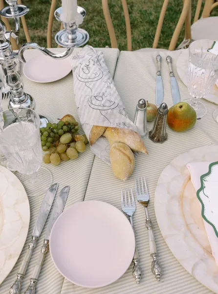Reception tablescape with an outdoor reception table set in blush and white plates, crystal goblets, silver candelabra, and fruit on a striped cloth