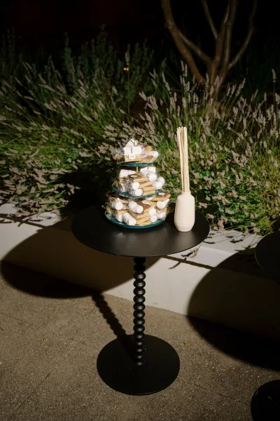 Wedding dessert table with a tiered dessert stand of wrapped cookies and marshmallows on a black pedestal table outdoors amid greenery