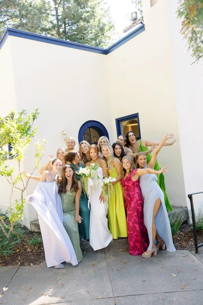 Bridesmaids group photo with bride with bridesmaids in colorful dresses holding bouquets outside a stucco building with arched doorway