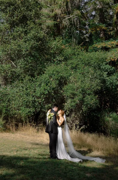 Wedding kiss portrait of bride and groom kissing, her long veil flowing as she holds a calla lily bouquet on a grassy field with trees