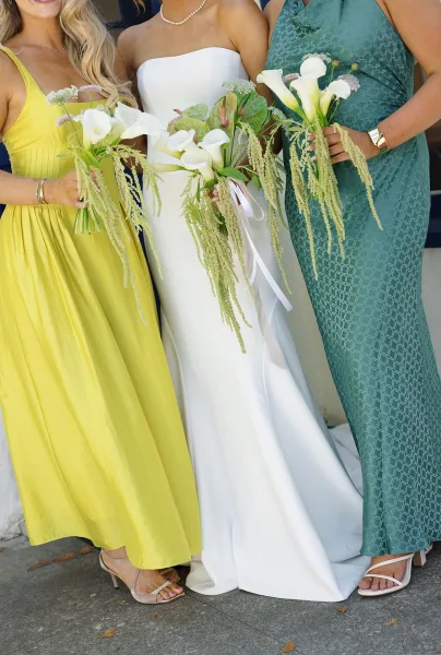 Bridesmaid bouquet of white calla lily bridesmaid bouquets with cascading greenery, white ribbon wrap, held by women in colorful dresses on stone pavement