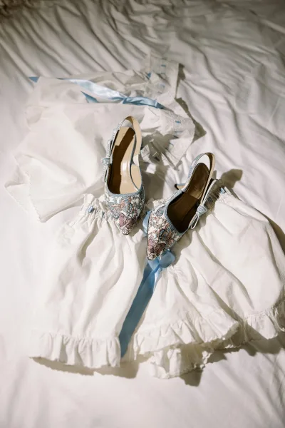 Wedding shoes and embroidered bridal heels with a blue ribbon beside white lace lingerie on crisp white bedsheets in a bedroom flatlay