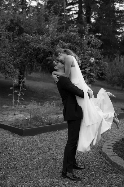 Wedding kiss portrait of groom lifting bride into a dip kiss, her veil flowing, on a garden lawn beside stone edging and a gravel path
