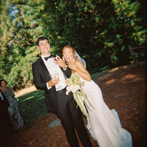 Couple portrait of bride and groom showing rings, holding champagne flutes, with veil and tropical bouquet on an outdoor path by evergreens