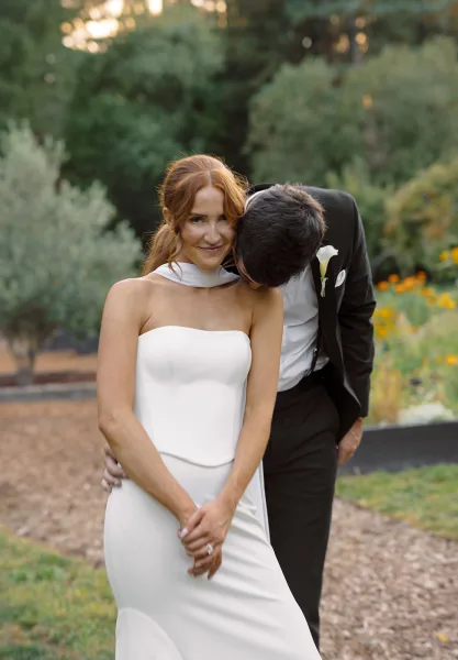 Couple portrait of groom kissing bride’s cheek as she looks at camera, showing her wedding ring on a gravel garden path