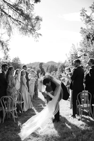 Wedding recessional dip kiss as newlyweds embrace in the aisle, her long veil flowing while guests cheer on a sunny outdoor lawn