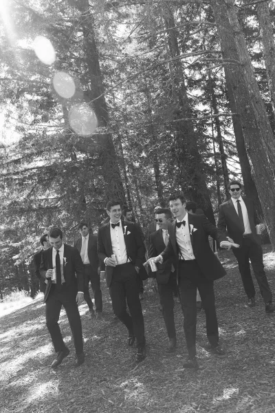 Groomsmen portrait of men in black tuxedos and bow ties wearing sunglasses, holding beverage cans in a sunlit pine forest clearing