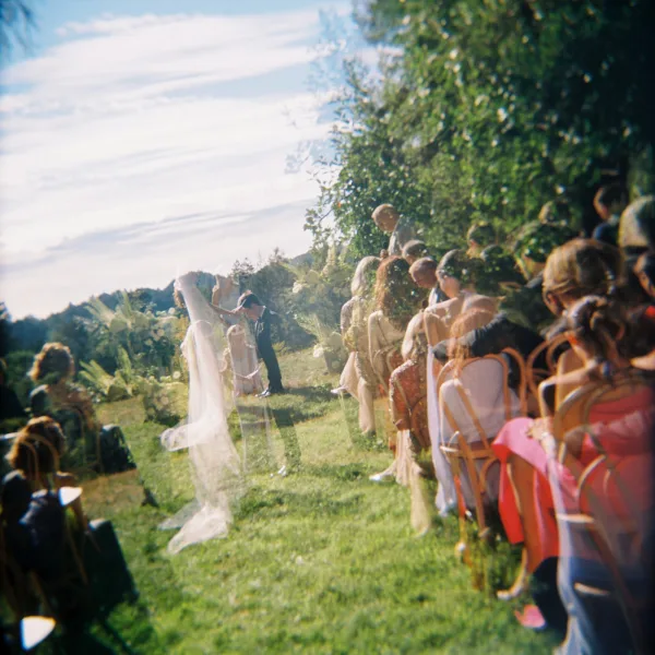 Ceremony moment at outdoor wedding ceremony as bride and groom stand at a floral arch, veil drifting on a sunny hillside lawn