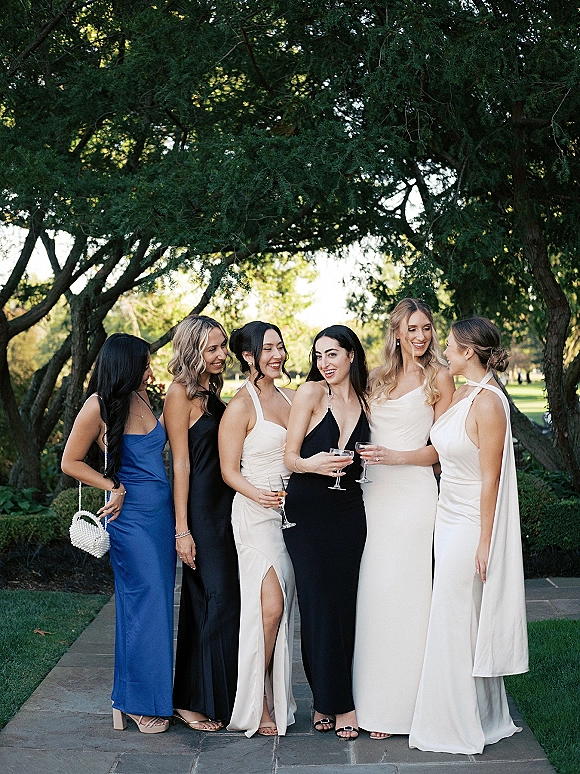 Bridesmaid group photo of friends holding champagne in mismatched black, white, and blue dresses on a garden stone pathway, pearl purse accent
