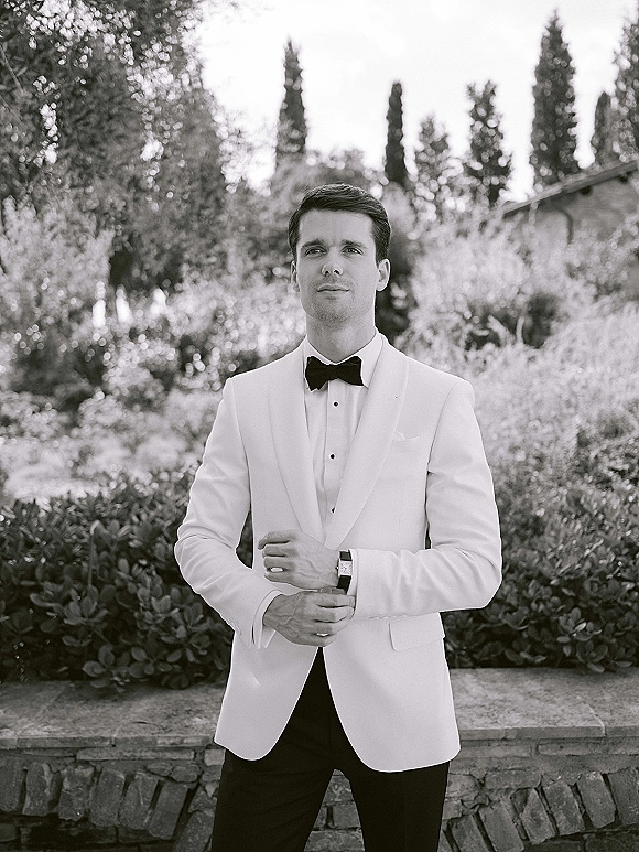 Groom portrait in a white tuxedo with black bow tie, standing by garden foliage and tall trees near a stone wall and roofline