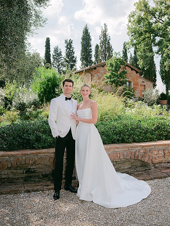 Couple portrait of bride and groom posing on a gravel garden path, bride in strapless gown with train, groom in white jacket by brick wall