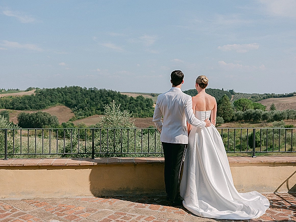 Couple portrait from behind of bride in strapless gown with long train and groom in white tuxedo jacket on terrace overlooking hills