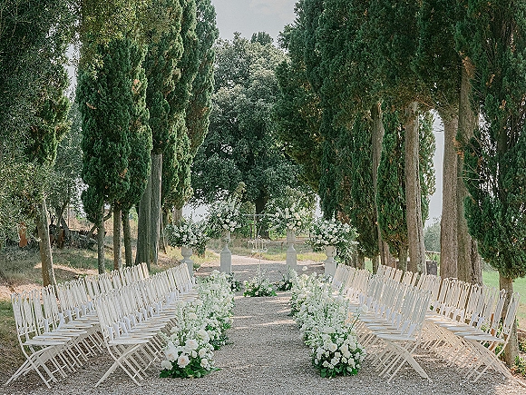 Outdoor ceremony setup with garden wedding ceremony rows of white folding chairs along a gravel aisle, lush greenery and metal arch under evergreens