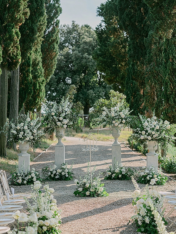 Ceremony altar decor with white floral arrangements and greenery on stone pedestals, framing a gravel garden ceremony aisle with chairs