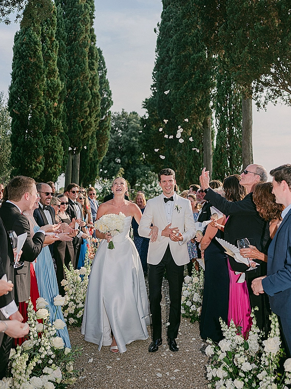 Wedding recessional as bride and groom walking aisle while guests toss rose petals, bride with white bouquet on a tree-lined gravel path