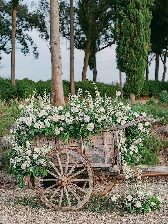 Floral cart decor with a rustic flower cart draped in white roses, greenery garland, and trailing blooms on a gravel garden path