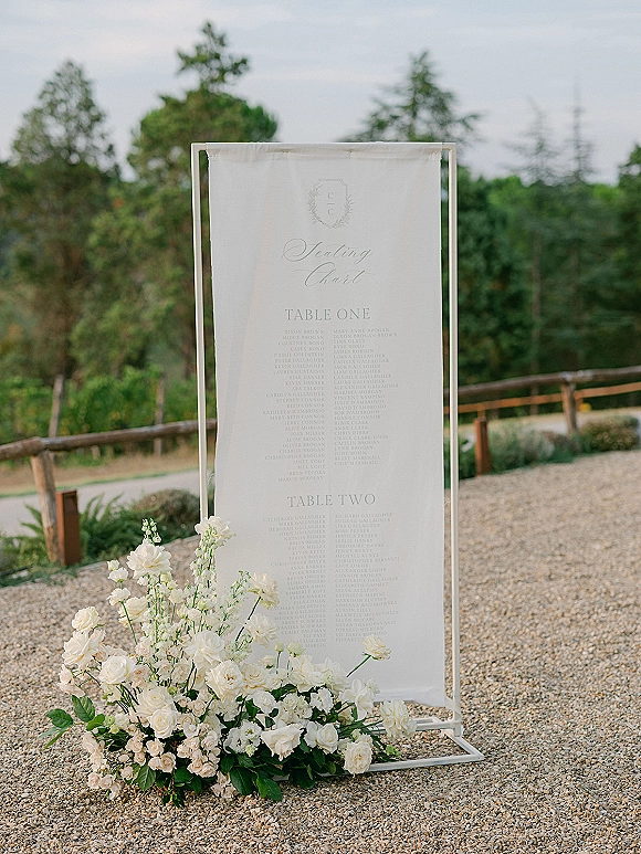 Wedding seating chart acrylic seating chart sign in a clear frame with white fabric panel and rose greenery base on a gravel path outdoors