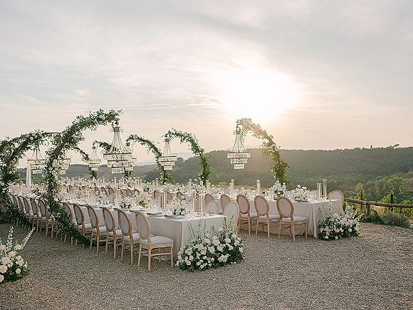 Reception tablescape with long banquet tables, white linens, greenery garlands and candlelight beneath crystal chandeliers on a sunset terrace