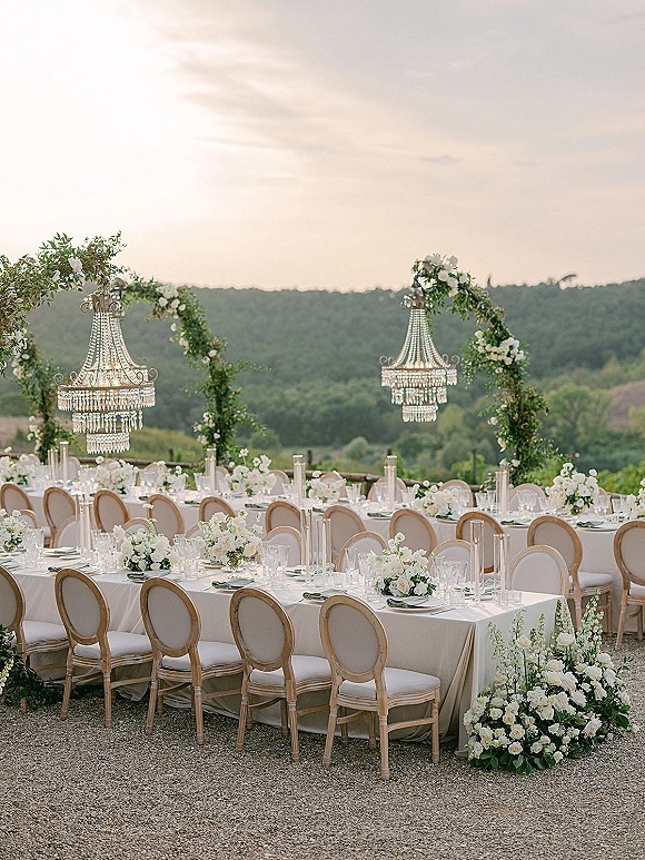 Outdoor reception tablescape with long banquet table wedding details, white linens, crystal chandeliers, greenery arches, mountain backdrop