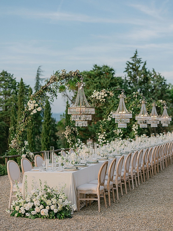 Reception tablescape for an outdoor wedding reception with a long banquet table, white roses and greenery garland, taper candles, and crystal chandeliers in a garden setting