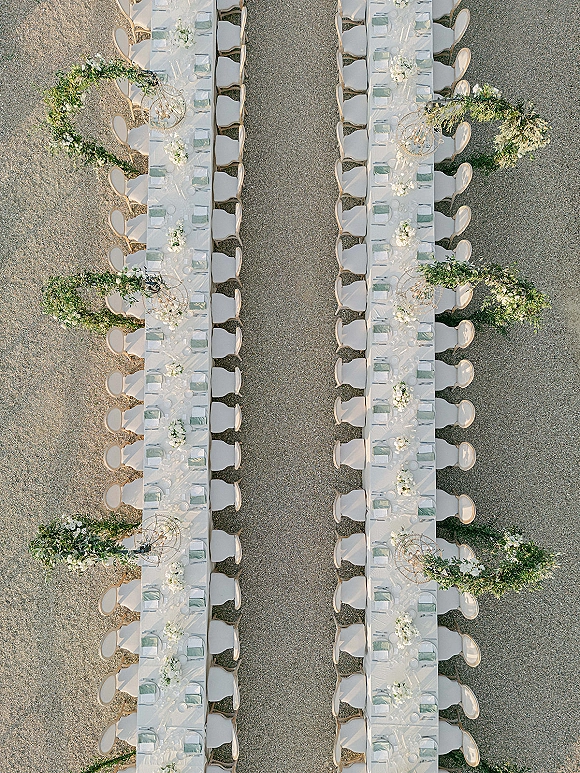 Reception tablescape with long banquet tables, white linens, floral centerpieces, and greenery garlands under chandeliers in a gravel courtyard