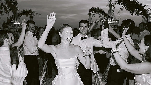 Wedding reception dancing with bride and groom in tuxedo and wedding dress, cheering with guests under chandelier lanterns outdoors at night