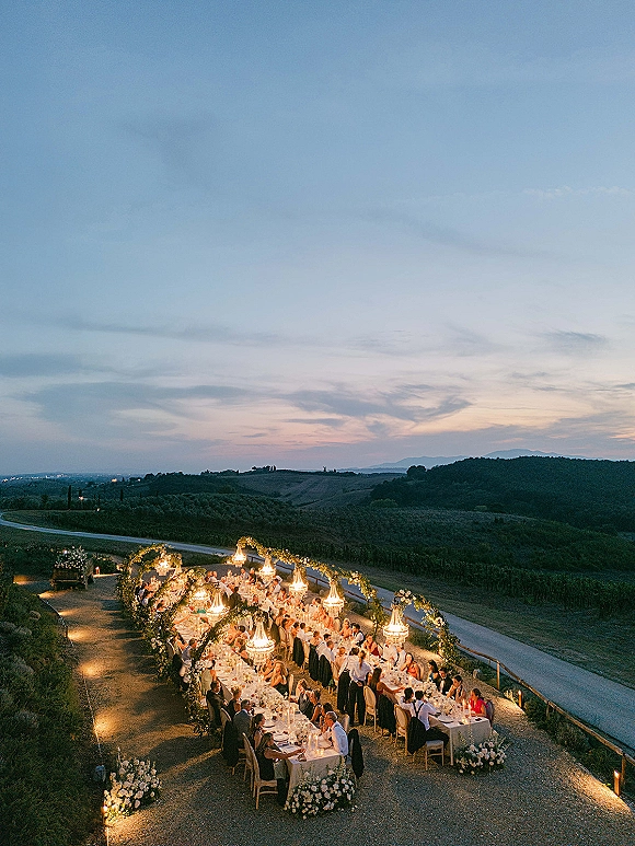 Outdoor reception setup with long banquet tables, white linens, and chandeliers glowing over candlelit florals in a vineyard at dusk
