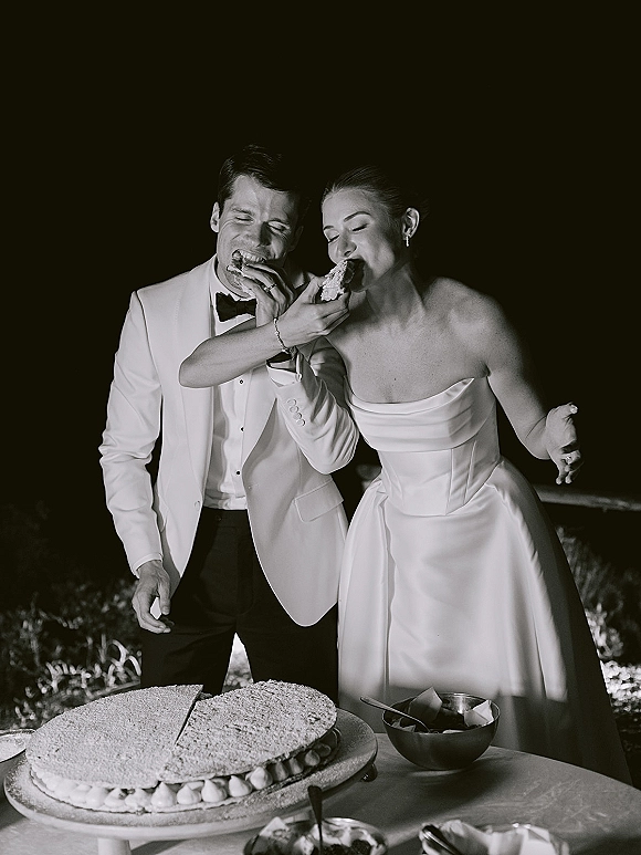 Wedding cake cutting as bride in a strapless dress and groom in a tuxedo share slices at an outdoor dessert table under night sky