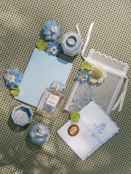 Wedding flat lay with wedding detail flat lay featuring rings in boxes, blue stationery, perfume, lace pouch, and blue flowers on a woven mat