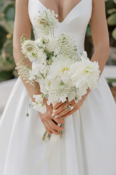 Bridal bouquet of white wedding bouquet blooms with greenery and ribbon wrap held by bride in V-neck gown, engagement ring, blurred foliage backdrop