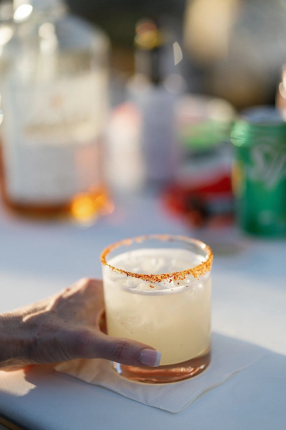 Wedding cocktail in a lowball glass with ice and a chili salt rim, held over a tabletop with cocktail napkin, bottles, and soda can