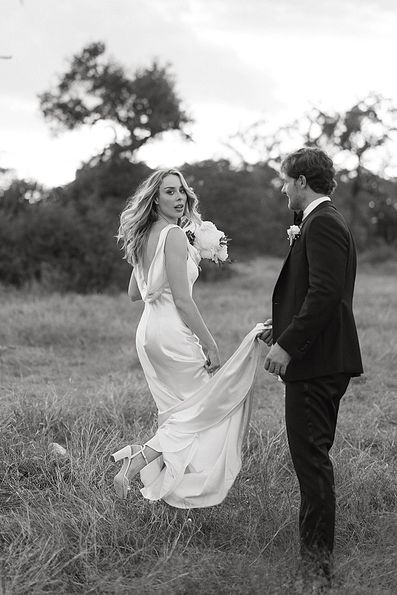 Couple portrait in black and white, bride looking back holding a bouquet as groom holds her dress train in a grassy field with trees
