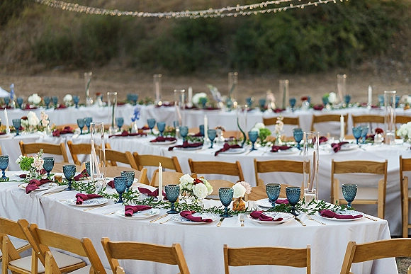 Reception tablescape for an outdoor wedding reception with long tables, blue goblets, burgundy napkins, candles, and floral garlands under string lights