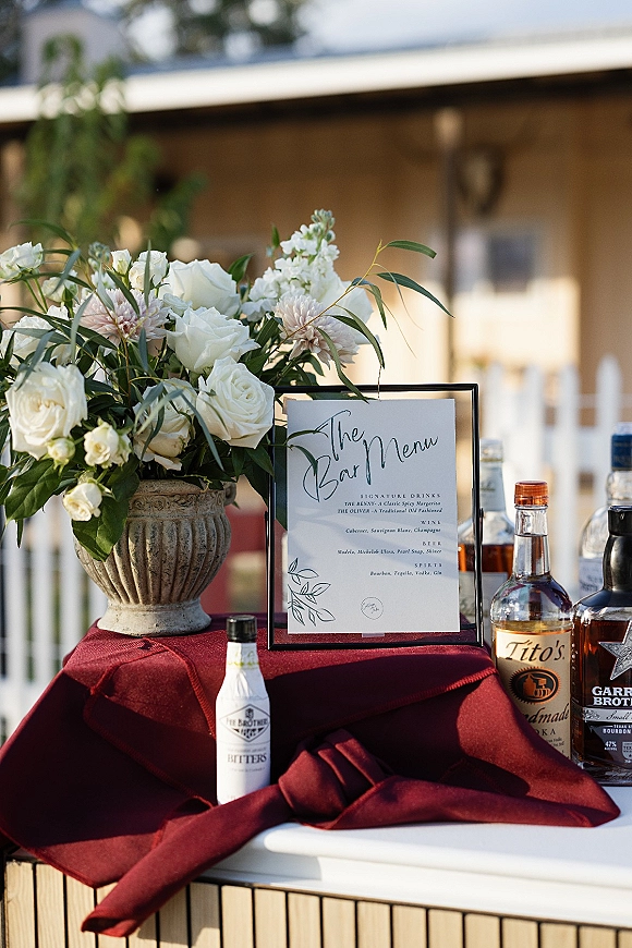 Wedding bar menu in a framed sign with white roses and greenery in a stone urn on burgundy linen, liquor bottles and bitters outdoors